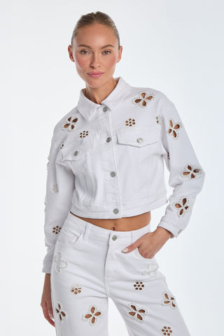 The woman wears a Kaia Pearl Jean Jacket - White, featuring a cropped silhouette and matching pants with embroidered beaded flowers. Her hair is tied back as she poses confidently against a plain background, hand on her hip.