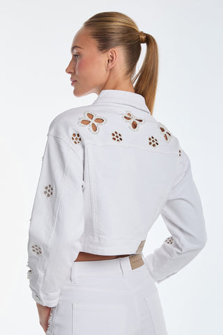 A person with a ponytail is wearing the Kaia Pearl Jean Jacket - White, showcasing its cropped silhouette and floral cutout designs on the back and sleeves against a plain background.