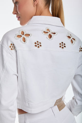 The woman wears a Kaia Pearl Jean Jacket - White featuring intricate cut-out designs on the back and shoulders, with embroidered beaded flowers. Her tied-back hair complements her white high-waisted pants, highlighting the jacket's elegance against the plain backdrop.