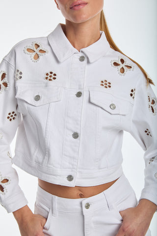 The woman wears a Kaia Pearl Jean Jacket in white with matching pants. The piece features floral cutouts and detailed brown crochet on the sleeves and front, forming an elegant outfit set against a simple background.