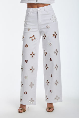 Dressed in the Kaia Pearl High Rise Jean - White, featuring a high waist, wide-leg cut, and embroidered beaded flowers with button and zipper closure. Paired with open-toed heels on a plain white background.