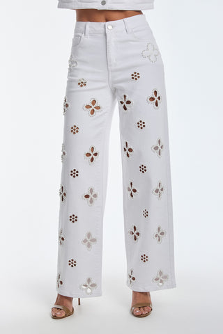 A person is wearing Kaia Pearl High Rise Jeans in white, featuring floral cutouts, paired with a matching crop top. The look is completed with open-toed heels against a plain white backdrop.