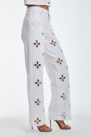 Someone in stylish white pants with floral cutouts, like the Kaia Pearl High Rise Jean - White, stands in heels. The detailed cutouts form a pattern along the leg against a plain, light-colored background.