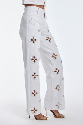 Side view of a person in the Kaia Pearl High Rise Jean - White, adorned with floral cutout patterns and embroidered beaded flowers. The stretchy jeans are paired with strappy heeled sandals against a plain white background.