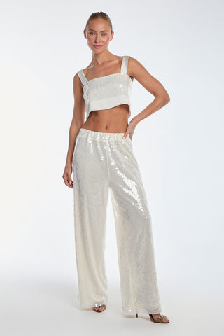 Wearing the Alba Sequin Pant in Pearl and a matching shimmering silver sequined crop top, a confident figure stands against a plain white background. With one hand on their hip and chic open-toed sandals, they capture the ideal resort wear look.