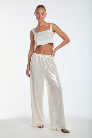 Wearing the Zoey Sequin Crop Top in Pearl, matched with wide-leg pants, a person confidently poses against a white backdrop. Their hair is neatly tied back as they stand with one hand on their hip, wearing open-toed shoes.