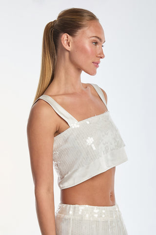 A woman with long blonde hair in a ponytail stands in profile against a plain background, wearing the Zoey Sequin Crop Top - Pearl, an elegant white sleeveless top with delicate sequin embellishments and a chic square neck design.