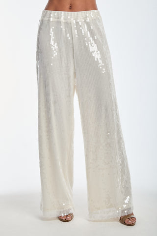 A person showcases the Alba Sequin Pant - Pearl, featuring high-waisted, wide-leg pants in a shimmering light color. Ideal for vacation wear, they have a reflective texture and are paired with open-toed sandals against a plain white background. Both feet of the wearer are visible.