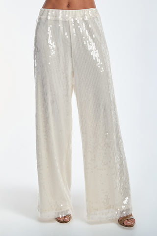 Someone is wearing the Alba Sequin Pant - Pearl, featuring wide legs, a high waist, and a stretch waistband. The light shimmering fabric reflects light brilliantly as they stand against a plain white background, revealing open-toed shoes.