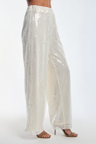 Dazzling in Alba Sequin Pant - Pearl, high-waisted with a cozy stretch waistband, they pair these shimmering wide-leg pants with elegant tan high-heeled sandals. The plain background enhances the stunning outfit.