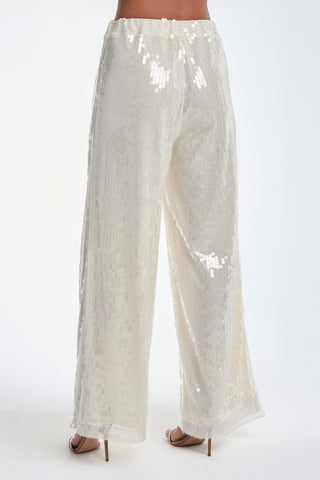 The person is wearing the Alba Sequin Pant - Pearl, featuring a high-waisted, wide-leg silhouette in a light hue. The sheer, flowy design evokes resort vibes. Beige high heels are visible as they stand against a plain white backdrop with their back to the camera.