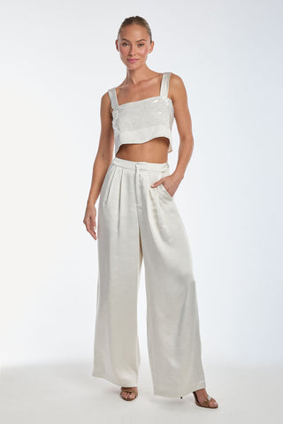 Modeling the Zoey Sequin Crop Top in pearl, paired with wide-legged satin trousers, a person poses against a white background. With one hand in their pocket and an elegant smile, they exude effortless charm.