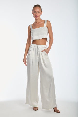 A woman stands confidently against a white background, wearing a stylish white sequined crop top and the wide-leg Valentina Satin Pant - Pearl. With her hair tied back, she smiles slightly with one hand in her pocket and the other by her side.