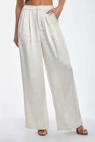 Someone is wearing the high-waisted Valentina Satin Pant in Pearl, displaying a wide-leg silhouette and pleats in a luxurious satiny fabric. Paired with open-toe sandals against a white backdrop, the elegance of the cream-colored ensemble is accentuated.