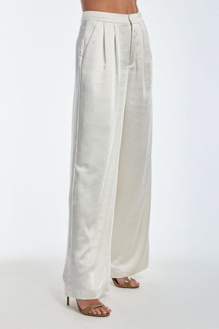 A person in high-waisted Valentina Satin Pants - Pearl, featuring a wide-leg design and pleated waist, stands against a plain white background. They wear open-toe high heels, and the pants showcase a smooth, silky texture.