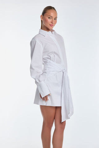 A woman is elegantly posed against a plain white background, wearing the chic Lana Wrap Dress - White with long sleeves and a knot at the waist. Her blonde hair is pulled back, complementing the classic shirt dressing style.