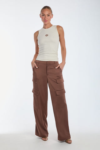 A person confidently poses with hands in pockets, wearing a white sleeveless top and Milan Satin Cargo Pant - Mocha, featuring intricate cargo detail, against a plain white background.