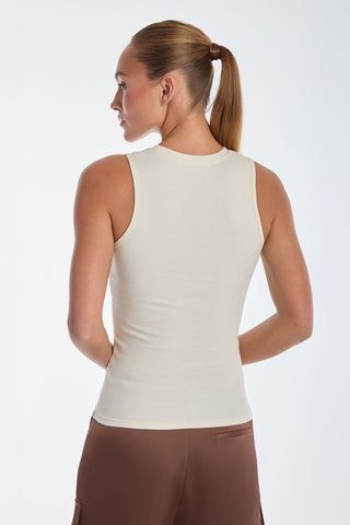 A person with a ponytail is wearing a sleeveless, ribbed top similar to the Rosette Tank Top - Pearl, paired with brown pants. They gaze slightly to the side against a plain white background.