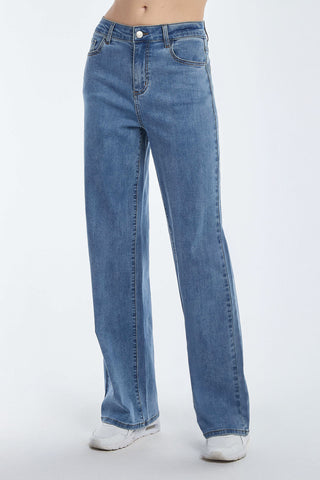 Wearing Dahlia High Rise Relaxed Jean in medium blue, a person stands with hands by their sides. They sport white sneakers, and the relaxed cut complements the plain, light-colored background perfectly.