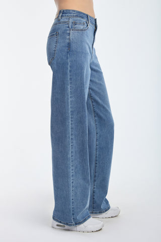 Side view of a person wearing Dahlia High Rise Relaxed Jean in medium blue with white sneakers against a plain background. The jeans feature a classic design, pockets, and a relaxed cut.