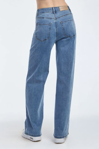 A person is shown from the waist down, wearing Dahlia High Rise Relaxed Jeans in Medium Blue with visible back pockets. They are barefoot on a plain white background.