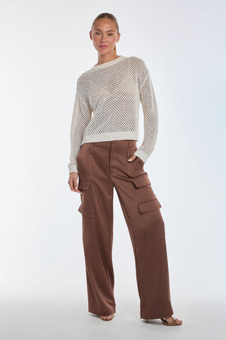 Wearing a resort-ready Lucie Knit Sequin Sweater - Pearl and brown cargo pants with large pockets, a person stands against a plain background with short hair tied back, posing with one hand in their pocket.