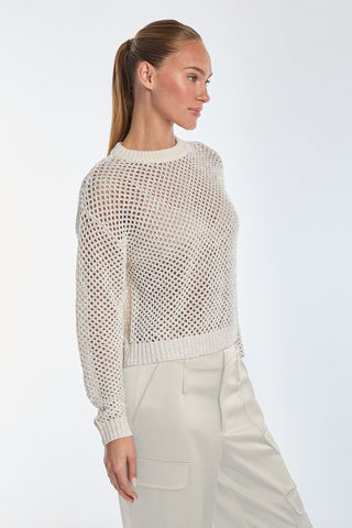 A person with long hair in a ponytail, radiating chic style, wears the Lucie Sequin Crochet Top - Pearl with long sleeves, paired with beige trousers against a white backdrop. The crocheted top elegantly exposes glimpses of a lighter undershirt beneath.