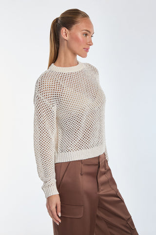 A person with long hair in a ponytail looks glamorous in their Lucie Knit Sequin Sweater - Pearl and brown pants. They stand sideways, hands in pockets, gazing to the side against a plain white background.