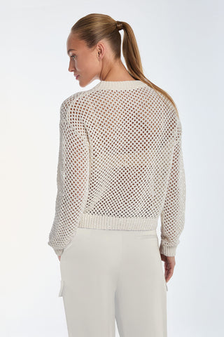 A woman with a ponytail embodies chic style in a white, sheer knitted sweater and pants, capturing the essence of the Lucie Sequin Crochet Top - Pearl as she gazes left against a light background.