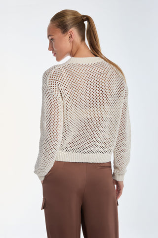 With long hair in a ponytail, they don a glamorous Lucie Knit Sequin Sweater - Pearl and brown pants. Standing against a light background, they exude a resort-ready vibe while looking to the side.