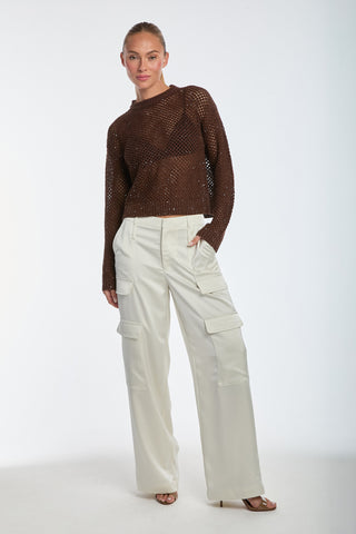 A woman in a brown knitted long-sleeve sweater and Milan Satin Cargo Pant - Pearl stands against a plain background. Her hair is pulled back, with one hand in her pant pocket, paired with heeled sandals.