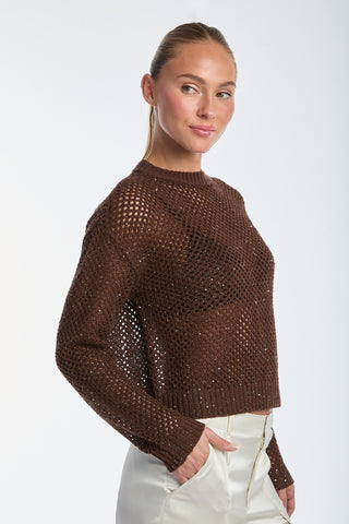 Wearing the Lucie Knit Sequin Sweater in Espresso and white high-waisted pants, a person poses chicly against a plain background. The textured open-weave design and their slight smile create a perfect resort-ready look as they gaze to the side.