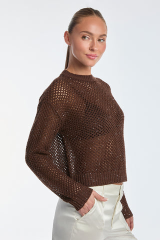 A person wearing the resort-ready Lucie Sequin Crochet Top in Espresso and white pants stands against a white background, looking to the side with a subtle smile and neatly pulled-back hair.