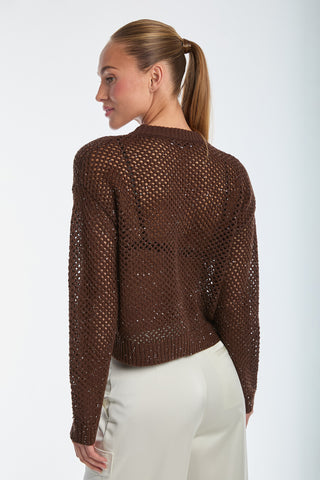 With her slicked-back ponytail, she exudes chic style in the Lucie Knit Sequin Sweater - Espresso and beige pants. The textured open-knit design of the sweater perfectly complements her resort-ready look against a plain white background.