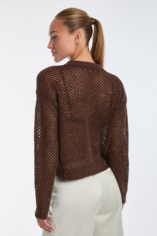 A person with long hair in a ponytail is wearing the glamorous Lucie Sequin Crochet Top in Espresso and off-white pants, with a textured, open stitch pattern. They are facing slightly away against a plain white background.