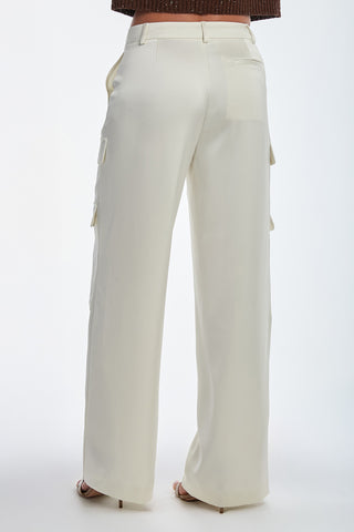 A person models the Milan Satin Cargo Pant - Pearl against a plain white background, showcasing its high-waisted, wide-legged design and side pockets. This cream color complements a dark top and high heels, offering an elegant yet functional look.