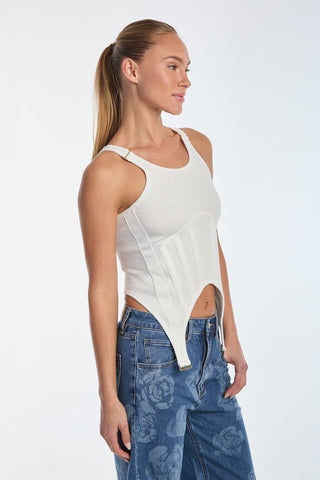 A person with long hair tied back is wearing a fitted white Kiara Garter Tank Top featuring rib fabric and geometric detailing, paired with rose-patterned blue jeans. They stand against a plain background, looking to the side.