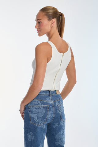 A person with a ponytail wears the Kiara Garter Tank in white, designed with ultra-soft rib fabric, corset-inspired lines, and a back zipper. They pair it with floral-patterned blue jeans while turned slightly to the side against a plain background.