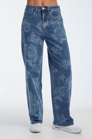 A person models Rosalie High Rise Relaxed Jeans in medium blue, showcasing a faded floral pattern with white sneakers against a white backdrop.