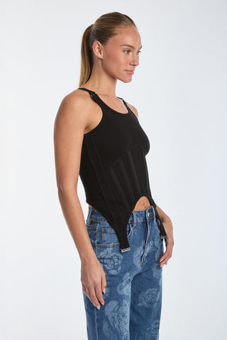 A person with long hair wears a Kiara Garter Tank Top - Black with corset-inspired details, paired with blue jeans featuring a floral pattern. They pose against a plain white background, looking to the side.