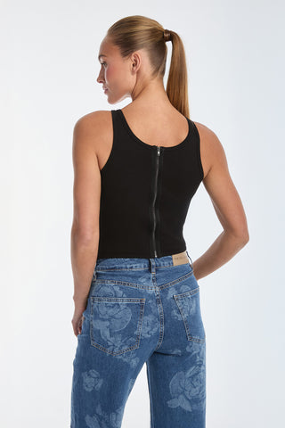 Dressed in a black Kiara Garter Tank Top, paired with floral-patterned blue jeans, they stand with one hand on their hip and look to the side, hair tied back in a ponytail. The corset-inspired design adds an edgy touch to the sleek ensemble.