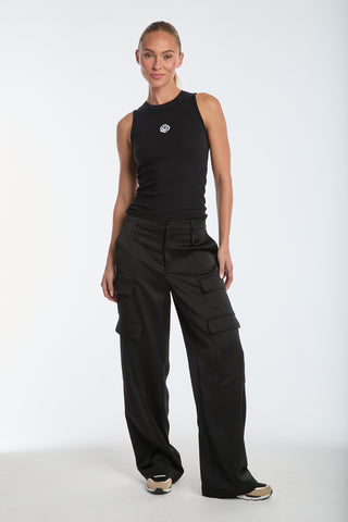 A person in a black Rosette Tank Top and black cargo pants stands against a white background. With sneakers on and sporting a short ponytail, they pose with one arm by their side and the other slightly bent.