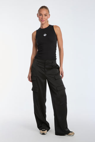 A person stands confidently against a plain background, wearing the Rosette Tank Top - Black, featuring a small circular logo and paired with black cargo pants. They complete the look with sneakers and an updo hairstyle.