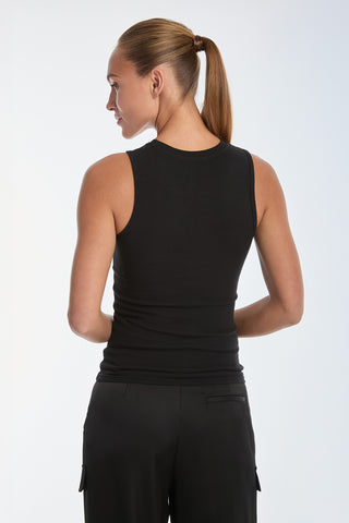 A brown-haired woman in a ponytail wears the black Rosette Tank Top and black pants, standing with her back to the camera in a softly lit studio.