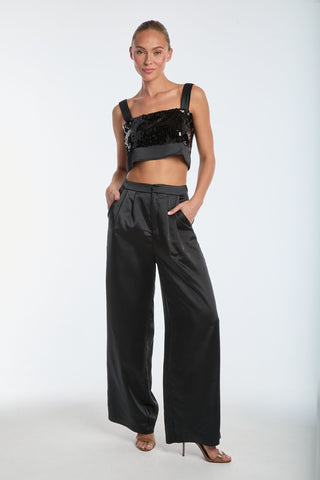 A model poses against a white background in strappy heels, wearing a black sequined crop top and Valentina Satin Pant - Black, featuring a high-waist wide-leg silhouette with hands in pockets.