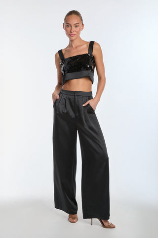 A person wearing a black sequined crop top and Valentina Satin Pant in black with a high-waist silhouette stands against a plain white background. They have their hands in pockets and are accessorized with high heels.