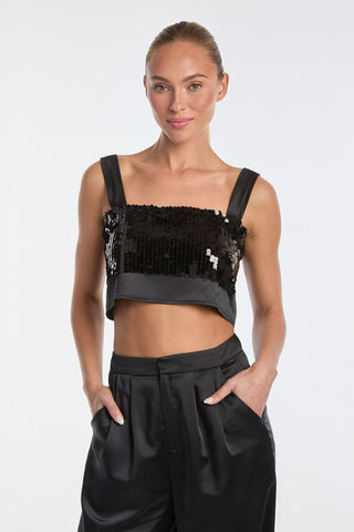 A person with tied-back hair confidently stands against a plain background, wearing the shimmering Zoey Sequin Crop Top - Black. Paired with sleek black satin pants and hands casually in pockets, the ensemble exudes a refined yet eye-catching allure.