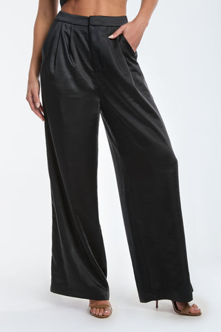 The image showcases a luxurious silhouette of the Valentina Satin Pant in black, featuring high waisted, wide-leg design with pleats, elegantly paired with high-heeled sandals against a plain background.