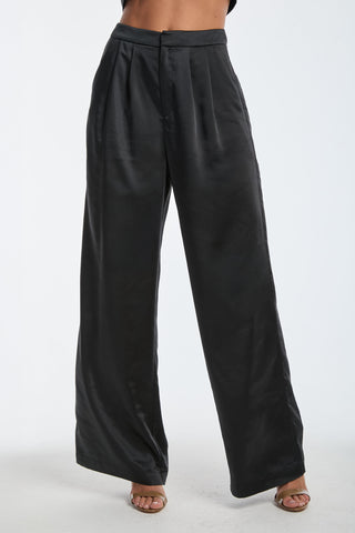 Wearing the Valentina Satin Pant in black, this person showcases high-waisted, wide-leg trousers with pleats and a button closure, paired with open-toe shoes, all set against a white background.