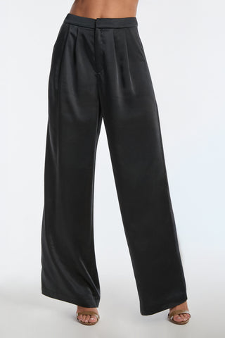 The Valentina Satin Pant - Black showcases a wide-leg silhouette crafted from sleek satiny fabric, complete with pleats and a high waist. Paired with open-toe heels, they create an elegant style against a plain background.